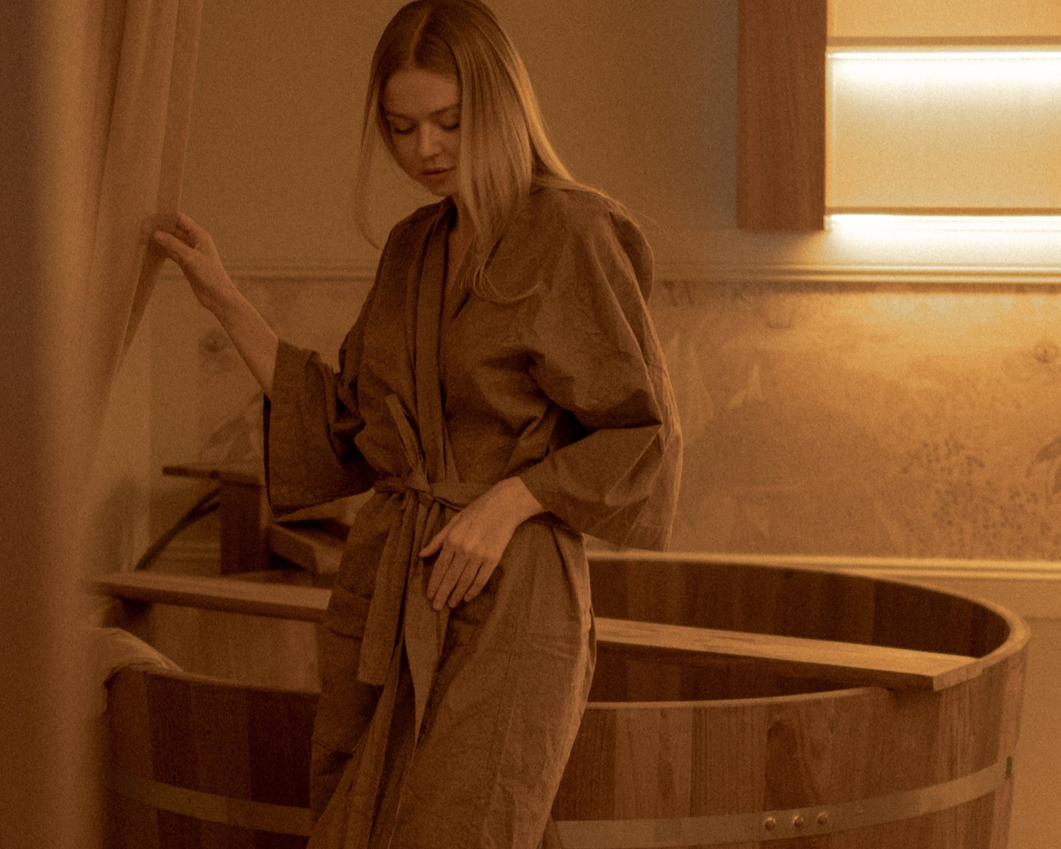 Woman in a robe standing next to a wooden bathtub in a warm-lit room.