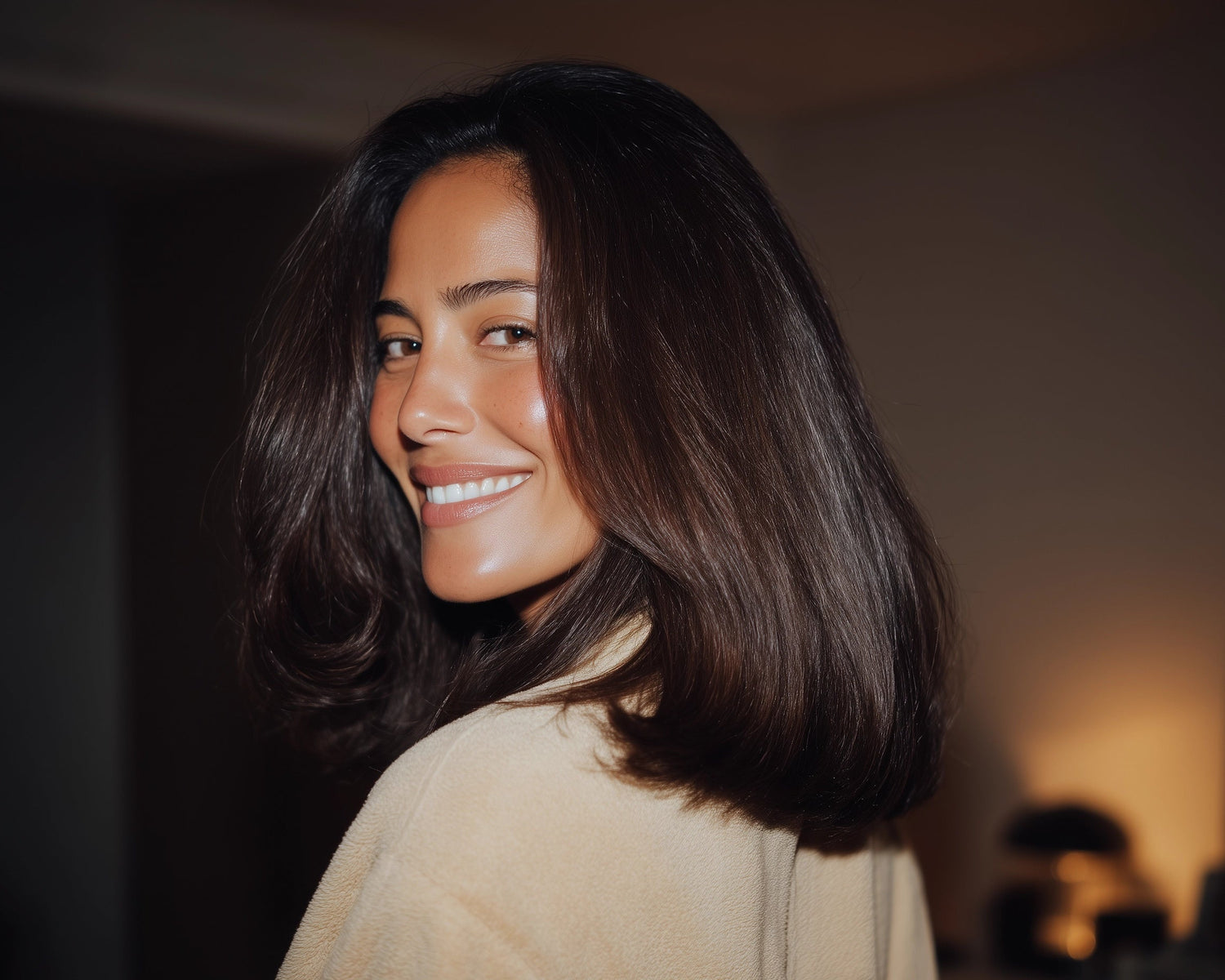 A woman with long, dark hair smiling over her shoulder, with a blurred background.